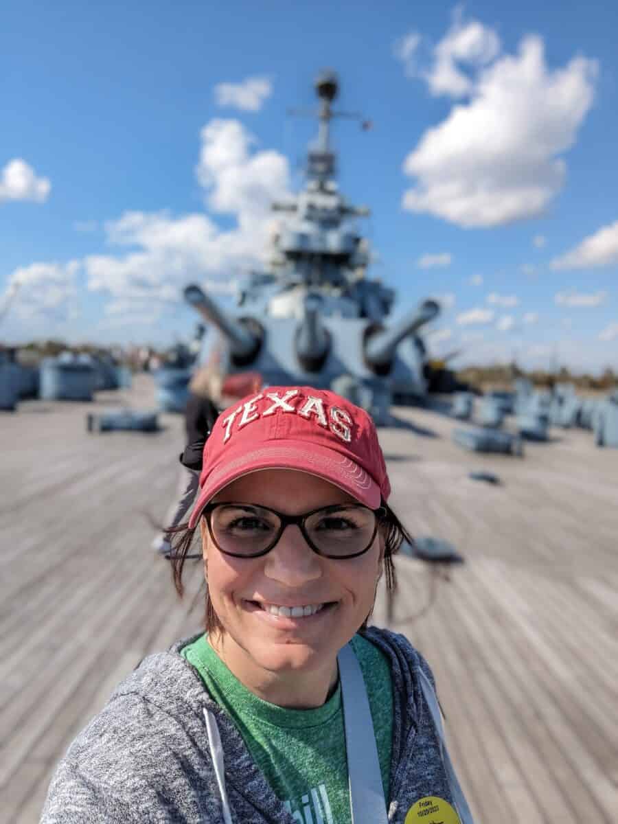 Selfie of Dusti standing on the top of the naval ship with guns in the background.